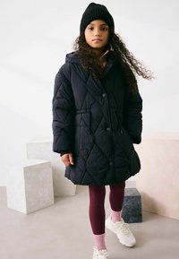 Must tikitud puffer-tüüpi mantel, millel on nööbitav kinnis ja kapuuts, kantud burgundia värvi soonikkoes retuuside ja valgete tennistega, millel on roosad sokid. Lokilised juuksed.