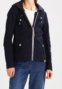 Woman wearing a navy hooded jacket with front zipper and floral-lined hood over a white top, paired with blue denim jeans.