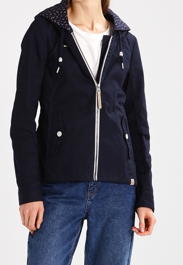 Woman wearing a navy hooded jacket with front zipper and floral-lined hood over a white top, paired with blue denim jeans.