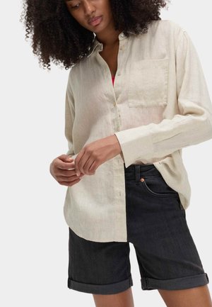 Jasnobeżowa lniana koszula z kieszenią i zapinanym na guziki kołnierzykiem, zestawiona z ciemnymi szortami dżinsowymi, z podwiniętymi mankietami. Skupienie na fakturze i dopasowaniu koszuli.