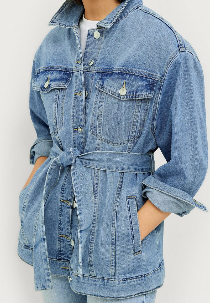 Ljusblå denimjacka med bälte i midjan, två bröstfickor och uppvikt ärm. Har en klassisk krage och knäppning.