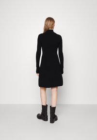 Frau mit langen Haaren trägt ein schwarzes, langärmeliges, knielanges Kleid und schwarze Stiefel bis zur Mitte der Wade, steht vor einem schlichten, hellen Hintergrund.