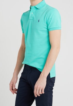 Mand iført en turkis kortærmet polo-shirt med logo og mørkeblå bukser, stående foran en ensfarvet hvid baggrund.