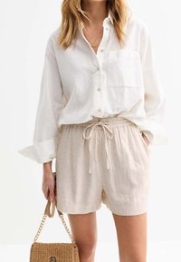 Žena v volné bílé košili na knoflíky a béžových šortkách na tkaničku, držící malou tkanou kabelku s řetízkem.
