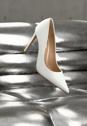 Zapato blanco de tacón alto con diseño de punta puntiaguda, superficie mate y lisa, y un interior nude, colocado sobre una superficie metálica brillante.