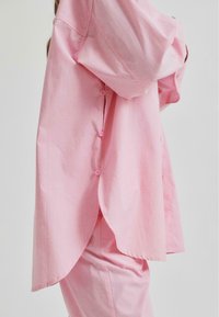 Camicia oversize rosa chiaro realizzata in tessuto morbido, con orlo curvo e chiusure a bottoni lungo il lato, design casual e confortevole.