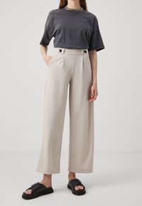 T-shirt gris oversize à manches courtes, pantalon beige à jambes larges avec poches latérales et accents de boutons, assortis à des sandales noires à lanières.