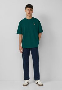 Groene katoenen t-shirt met korte mouwen, ronde hals en een klein logo op de linkerborst, gecombineerd met marineblauwe broek en witte sneakers.
