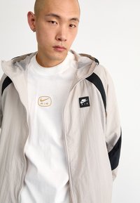 Nike windbreaker in light gray with black accents, featuring a zip front, mesh lining, and branded patches. Worn over a white tee.