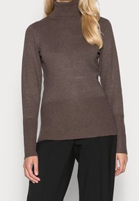 Femme portant un pull à col roulé côtelé marron à manches longues et un pantalon noir, vue du cou jusqu'aux hanches.