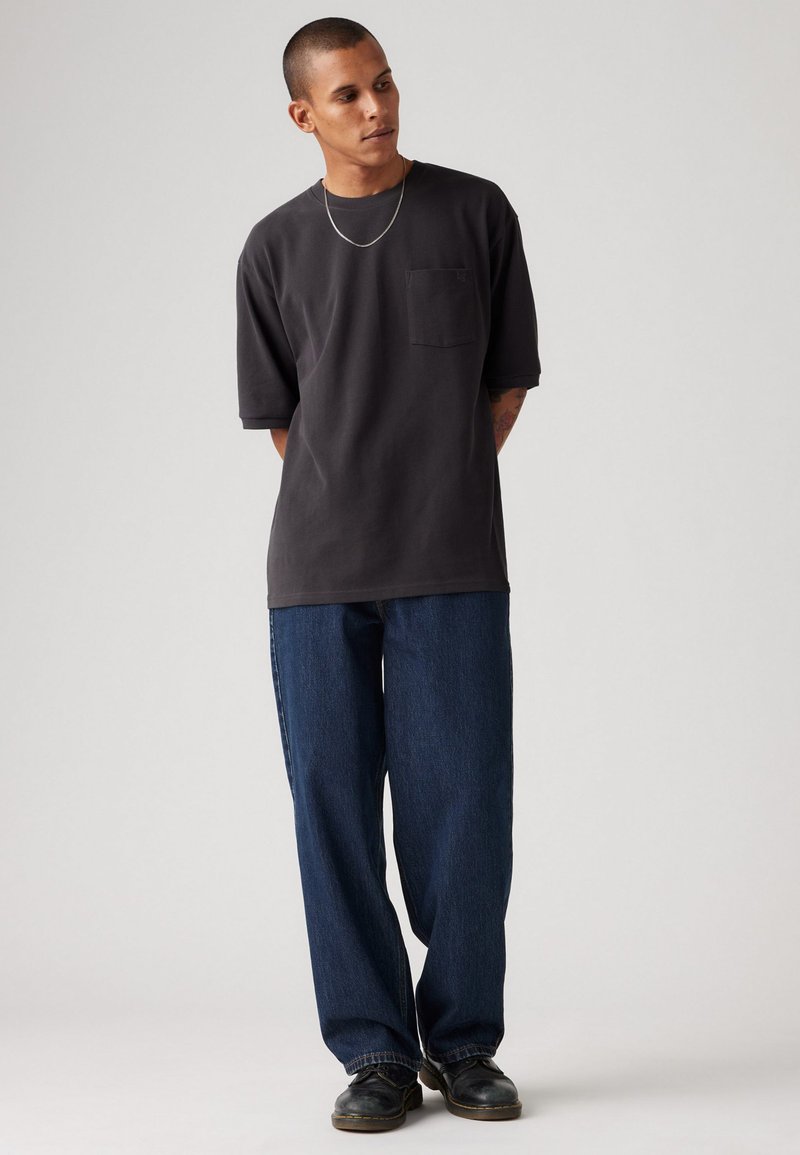 Dunkelgraues T-Shirt mit kurzen Ärmeln und einer Brusttasche, kombiniert mit locker sitzenden blauen Jeans und schwarzen Schuhen, vor einem einfarbigen Hintergrund.