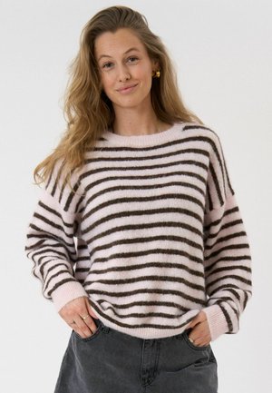 Naine, kellel on pikad lainelised juuksed, kuldsed rõngaskõrvarõngad ja roosa-pruunide triipudega kampsun, käed tumedates hallides teksapükstes taskutes.