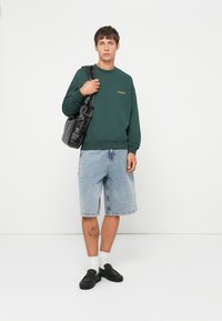 Green sweatshirt with orange logo, denim shorts with a faded look, black slip-on shoes, and a black shoulder bag. White socks visible.