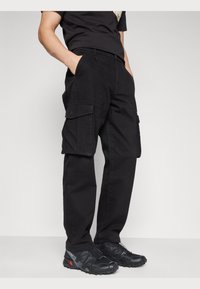 Black cargo pants made of cotton, featuring side pockets and a relaxed fit. Paired with black athletic shoes with textured soles.
