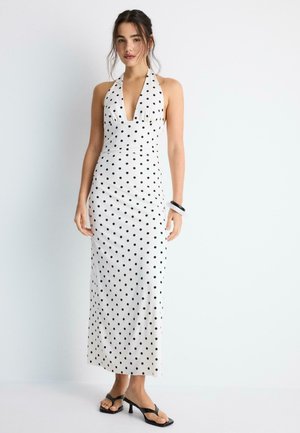 Frau trägt ein weißes Neckholder-Maxikleid mit schwarzen Polka-Dots und schwarzen Sandalen mit Absatz, stehend vor einem schlichten weißen Hintergrund.