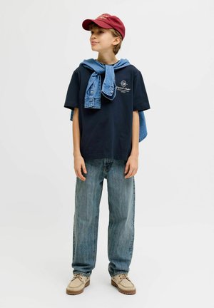 Jongen met rode pet, marineblauw T-shirt, spijkerjack om de schouders geknoopt, loszittende blauwe spijkerbroek en beige schoenen, staand tegen een effen achtergrond.