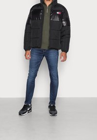 Chaqueta de abrigo negra con paneles de pecho brillantes, cierre de cremallera en la parte frontal y logo de Tommy Hilfiger. Lleva puesta sobre un forro polar verde, combinada con jeans ajustados azules y zapatillas negras.