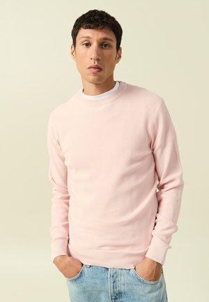 Mężczyzna o krótkich ciemnych włosach, ubrany w jasnoróżowy sweter z długim rękawem na białej koszuli i jasnoniebieskich dżinsach, stoi z rękami w kieszeniach.