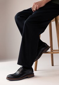 Black leather dress shoes with brogue detailing, a rounded toe, and wooden soles, paired with dark pants and a wooden stool backdrop.