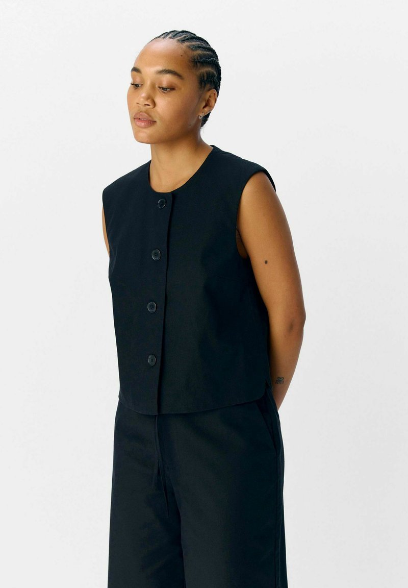 Zwarte mouwloze cropped vest met ronde hals, sluiting met knopen aan de voorkant en een gladde stoftextuur. Beschikt over zijnaadzakken en een getailleerd ontwerp.