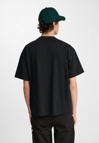 Zwart T-shirt met korte mouwen en een relaxte pasvorm, met een ronde halslijn. De persoon draagt een donkergroene pet, gezien vanaf de achterkant.