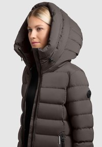 Puchowa brązowa kurtka z dużym kapturem, zapięciem na zamek z przodu oraz czarną naszywką z logo na rękawie. Gładka faktura, pikowany wzór.