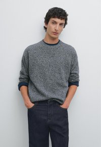 Grijze gebreide trui met een gestructureerd patroon, geribbelde marineblauwe manchetten, ronde halslijn, gecombineerd met donkere spijkerbroek. Eenvoudige lichte achtergrond.