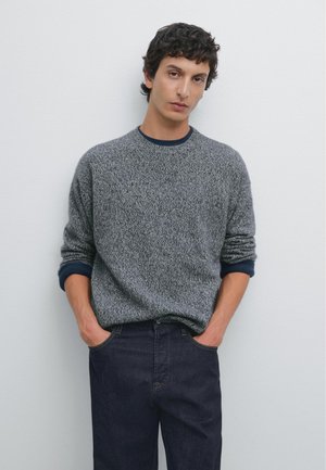 Maglione grigio a maglia con un motivo testurizzato, polsini a coste blu navy, scollo rotondo, abbinato a jeans in denim scuro. Sfondo chiaro e semplice.