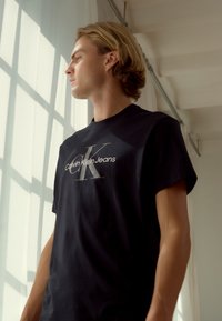 Camiseta de manga corta negra con un logo gris de 'Calvin Klein Jeans' y una gran 'CK' en una fuente minimalista. Textura de algodón suave.