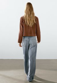 Hnědá semišová zkrácená bunda v kombinaci s světle modrými širokými džíny. Džíny mají zadní kapsy a volný střih. Model je viděn zezadu.