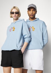 Lyse blå hettegensere med et tegneseriedesign foran, kombinert med svart og hvitt shorts. Modellen har på seg solbriller og en grå caps.