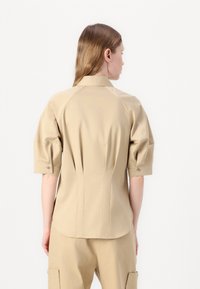 Camicetta beige con maniche corte e a sbuffo, dal design aderente. Tessuto liscio con piega sul retro e orlo dritto.
