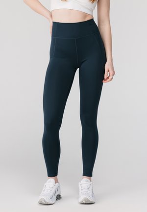 Kvinne iført svart høytlivslegging og hvite treningssko, stående med én hånd på hoften mot en ensfarget bakgrunn.