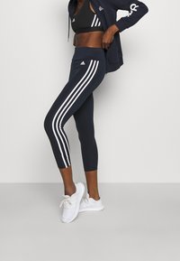 Marinblå träningstights med tre vita vertikala ränder, med hög midja. Kombineras med en svart sport-bh och vita sneakers.