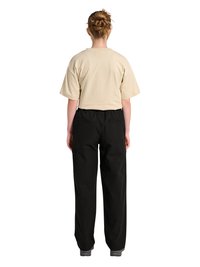 Persoană cu păr împletit poartă o cămașă bej cu mâneci scurte și pantaloni negri cu picioare largi, stând cu spatele la cameră pe fundal alb.