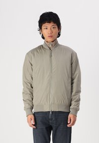 Giacca bomber leggera beige con colletto alto, chiusura a zip, polsini e vita a coste. Tessuto liscio con texture minimale.