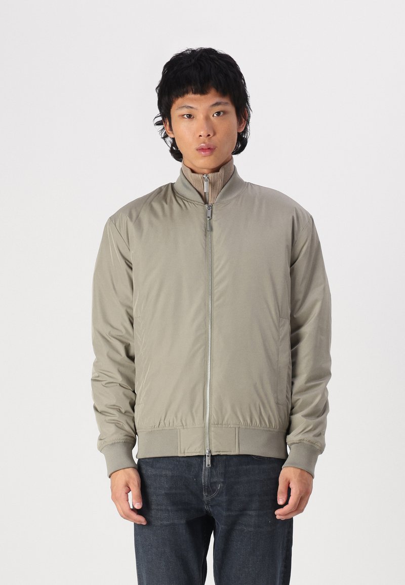 Giacca bomber leggera beige con colletto alto, chiusura a zip, polsini e vita a coste. Tessuto liscio con texture minimale.
