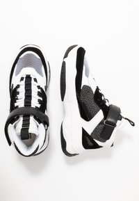 Černobílé tenisky z materiálů síťoviny a semiše, s silnou podrážkou, nastavitelným pásem a texturovanými akcenty.