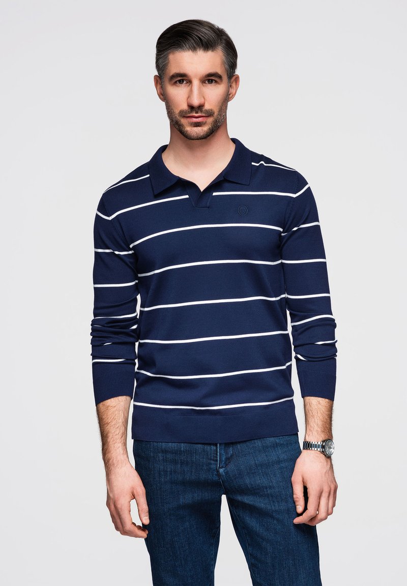 Mann trägt ein dunkelblaues Langarm-Poloshirt mit weißen horizontalen Streifen und blaue Jeans, steht vor einem einfarbigen hellen Hintergrund.