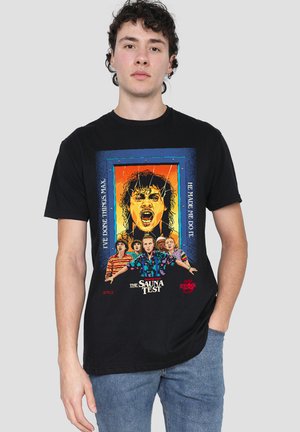 Young man wearing black graphic t-shirt featuring characters and text from Netflix's Stranger Things Chapter Four: The Sauna Test.