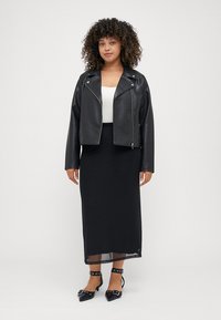 Black leather biker jacket, white top, black maxi skirt, and black shoes with straps. Curly hair frames the face, centered pose.
