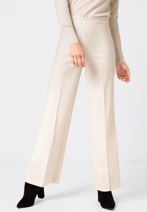 Vrouw draagt een hoge taille, wijd uitlopende beige broek en een bijpassende beige top met lange mouwen, gecombineerd met zwarte enkellaarzen.