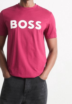 Person trägt ein rotes T-Shirt mit dem Wort "BOSS" in großen weißen Buchstaben, Hände in dunklen Hosentaschen, einfarbiger weißer Hintergrund.