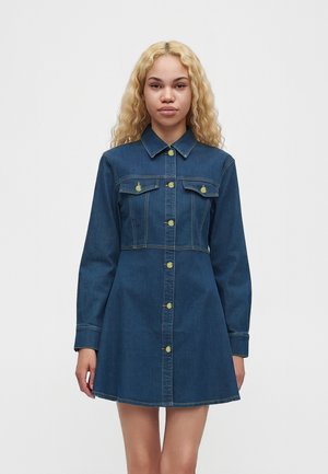 Ung kvinde med langt blondt hår iført en blå knaplukket denimkjole med lange ærmer og lommer på en hvid baggrund.