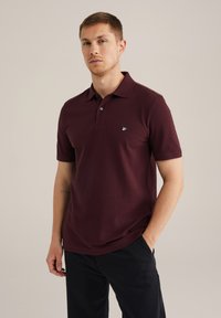 Burgundin värinen polo-paita klassisella kauluksella, lyhyillä hihoilla, kahden napin kädellä ja pienellä vihreällä emblemillä vasemmassa rinnassa. Venytettävää puuvillamateriaalia.