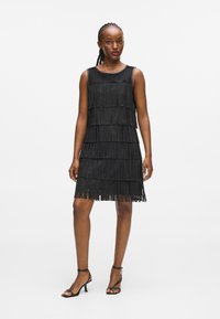 Fekete ujjatlan ruha réteges bojt részletezéssel, sima selyem felső résszel és térd hosszúságú szoknyával. Fekete magas sarkú cipővel párosítva.