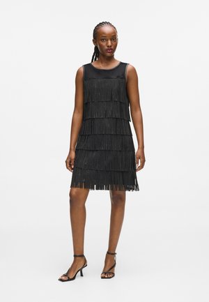 Robe noire sans manches avec des détails en franges superposées, haut en satin lisse et ourlet au genou. Associée à des talons hauts noirs.