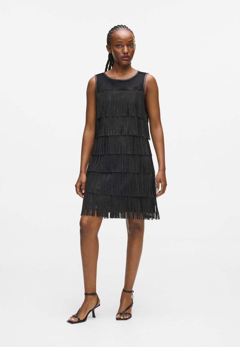 Fekete ujjatlan ruha réteges bojt részletezéssel, sima selyem felső résszel és térd hosszúságú szoknyával. Fekete magas sarkú cipővel párosítva.