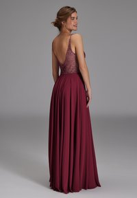 Joven mujer con un vestido de noche burdeos sin mangas, con cuerpo decorado con cuentas y espalda baja, de pie y sonriendo sobre un fondo gris liso.