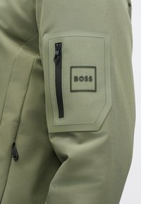 Khaki green jacket featuring a zippered pocket with a black accent and embossed "BOSS" logo. Smooth texture with a structured design.